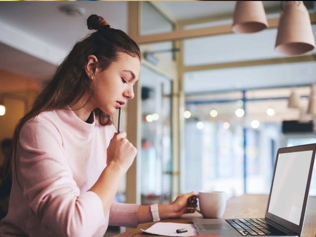 A girl sits at her laptop, sipping a cup of coffee, deep in thought as she brainstorms ideas for her essay.