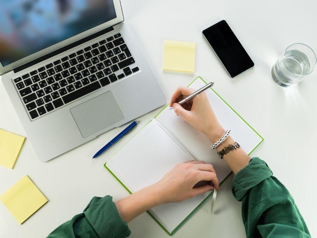 a girl writing on a notebook while her laptop and her phone lies beside her