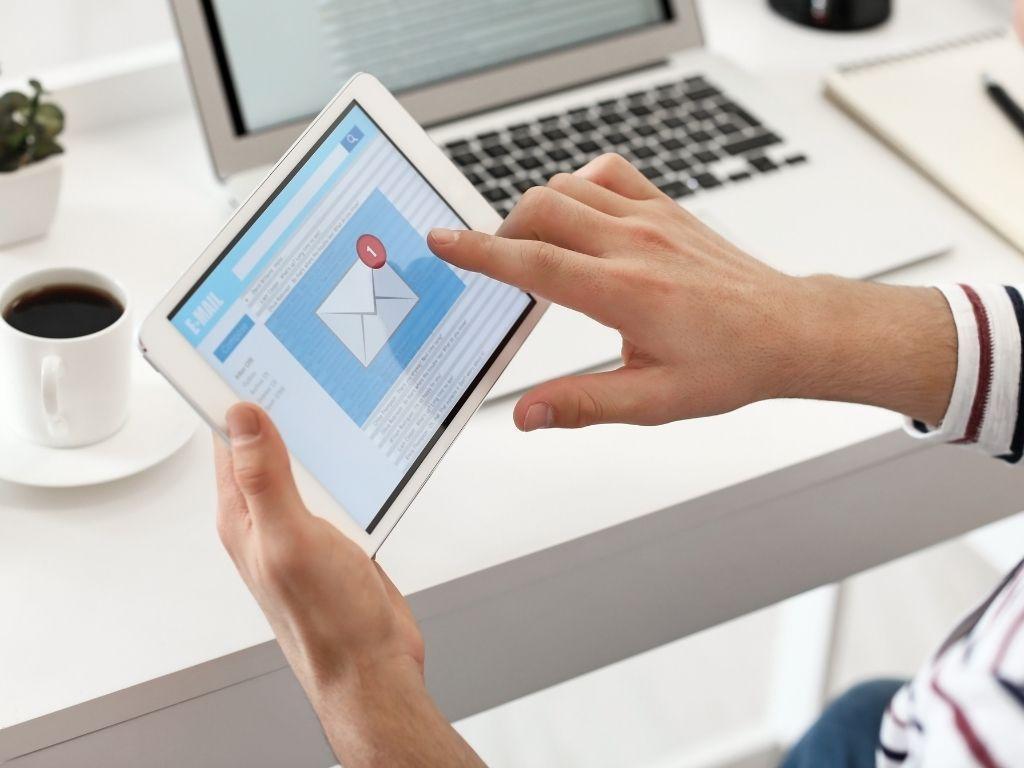 A person opening an email on a tablet, with a laptop and a cup of coffee resting on the table.