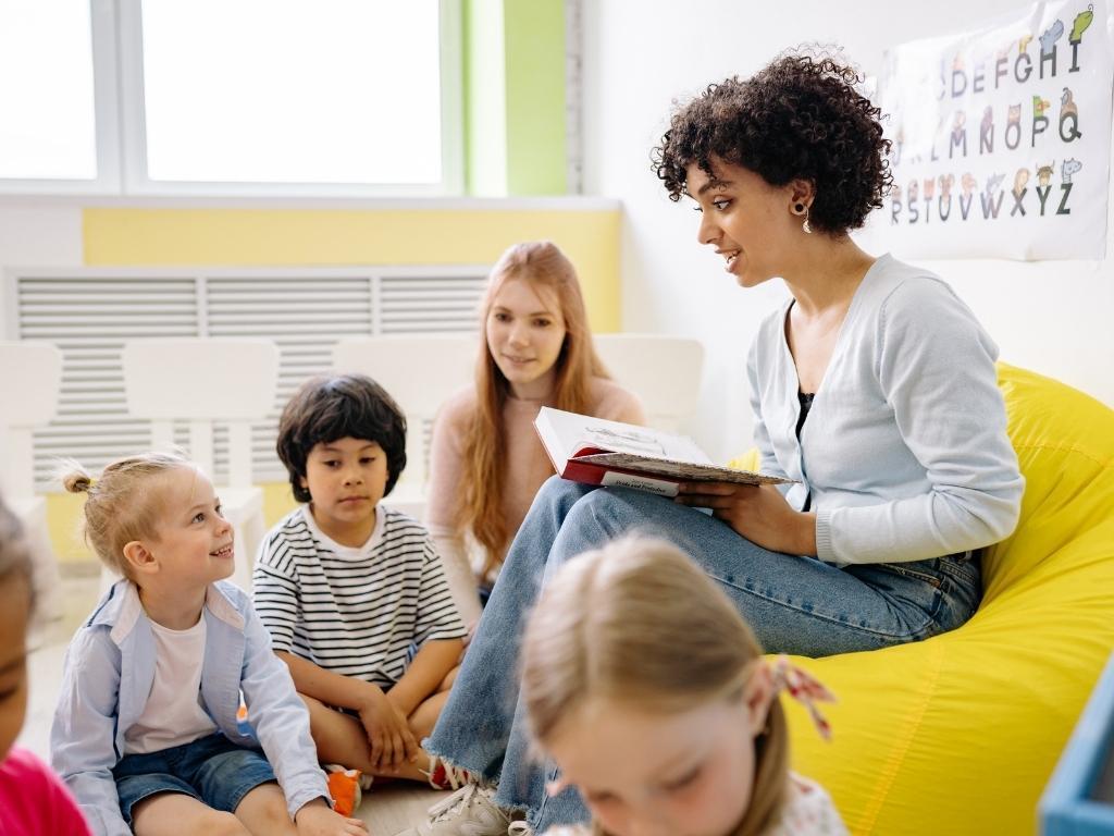 A female teacher reading a storybook to a group of kids, all smiling and enjoying the moment.