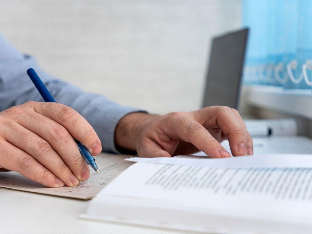 a man writing something with his pen while a book and his laptop is placed nearby