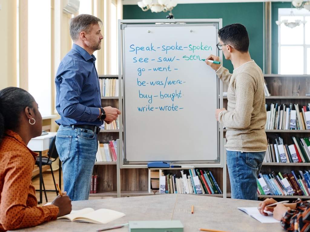 A man writing the forms of verbs on a whiteboard, while a man and a woman watch attentively.