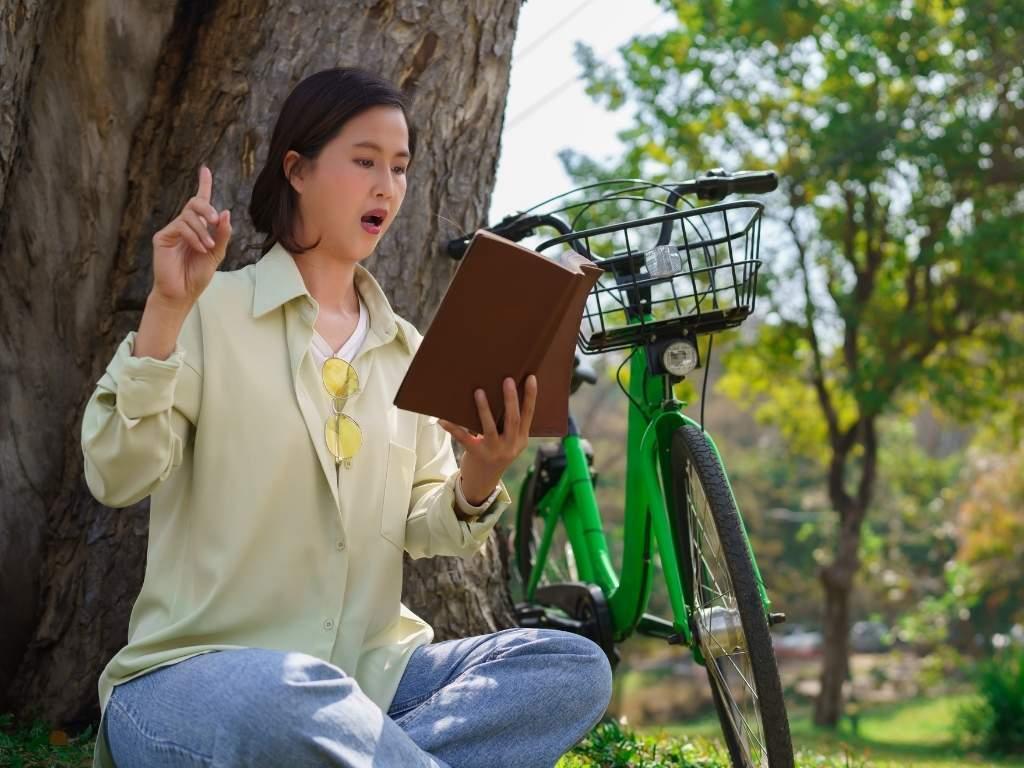 A gir reading from her book practicing on how to become an audiobook narrator while her bicycle is standing nearby