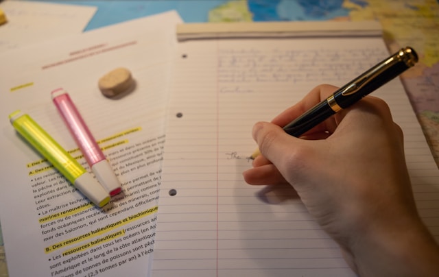 A person writing with a pen and with highlighters next to the book