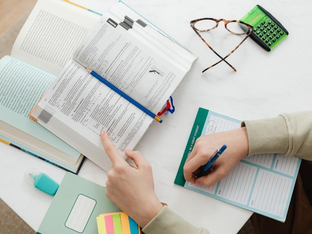 A person writing a report, with two books, a pair of glasses, and a calculator placed nearby on the table.