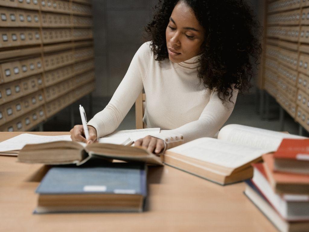 a girl writing on a notebook in a library with several books placed on a table.