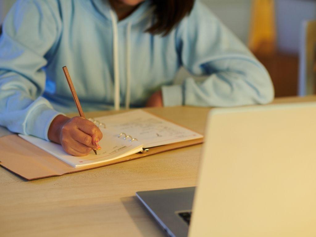 A girl writing on her notebook while her laptop is placed in front of her