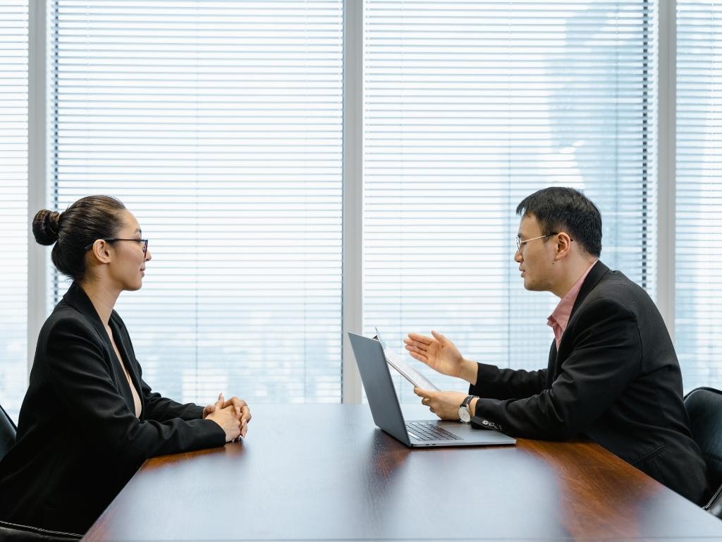 A man conducting an interview with a female, holding her CV in his hands, with a laptop placed nearby on the table.






