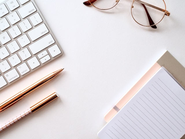 A table with a computer, pens, glasses, and notepad