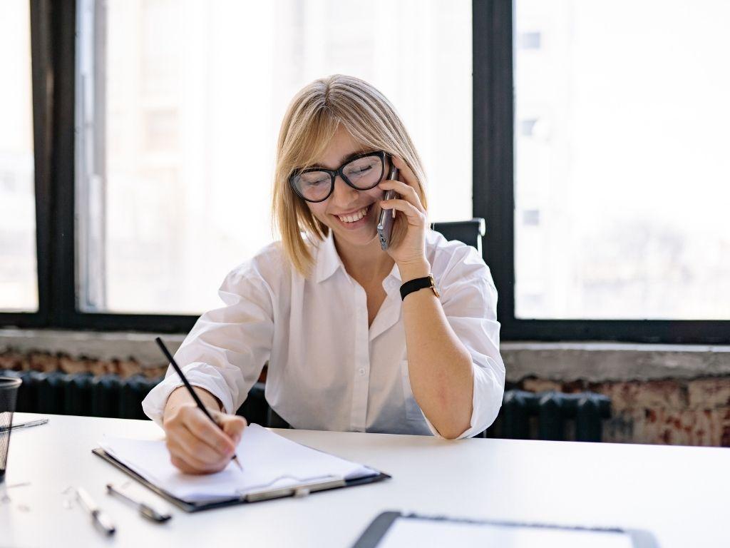 A girl on a phone call, filling out a form with a pencil, while sitting at a table.