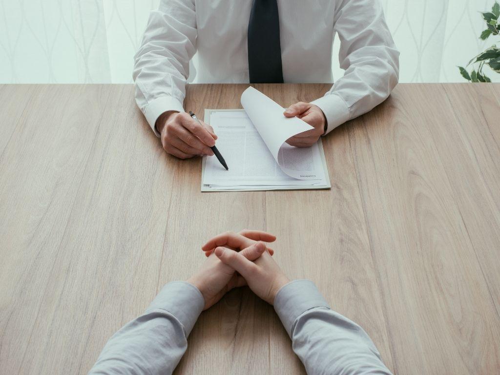 Two people sitting at a desk, one holding papers and seemingly conducting the interview.