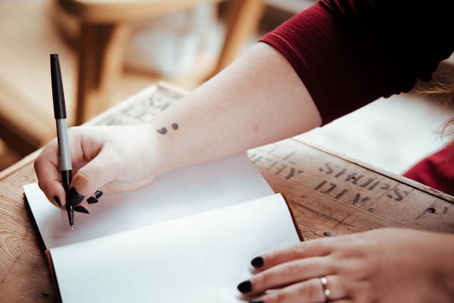 A person with arm tattoos writing in a notebook 