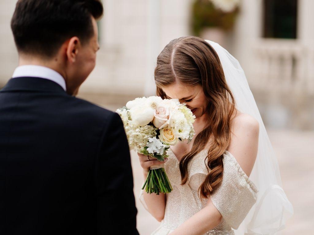 A bride holding a bouquet of flowers and smiling radiantly, while her groom looks at her with a loving smile on their wedding day.