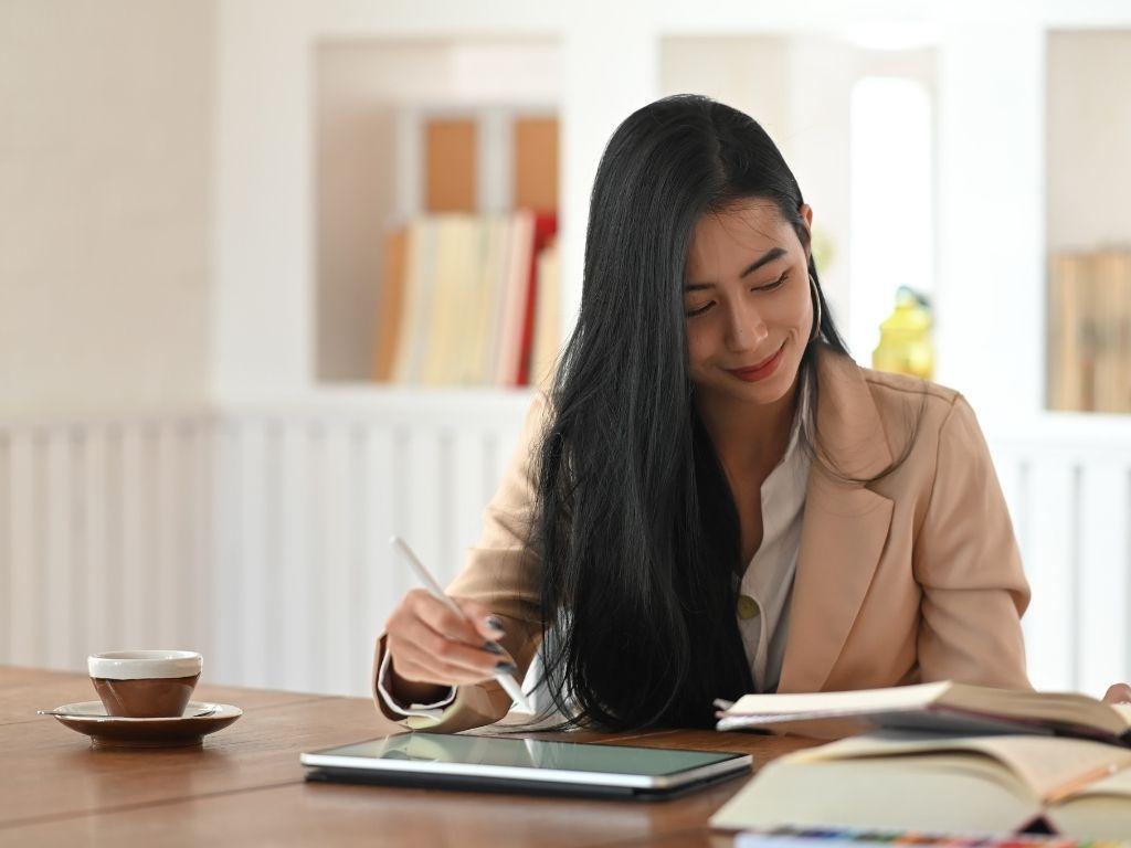 A girl working on her ipad while reading a book and another book placed nearby. 