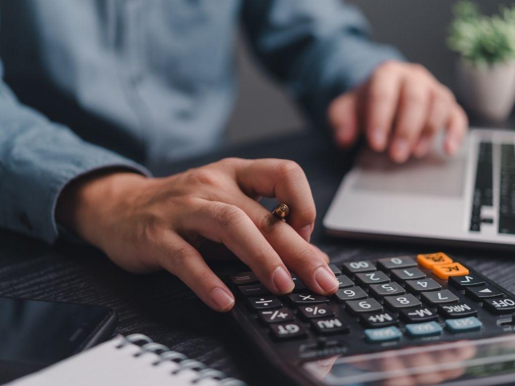 A man working on his laptop while using a calculator to perform calculations.