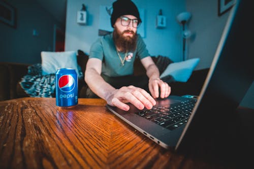 Free Bearded man wearing beanie, typing on laptop with soda can in cozy workspace. Stock Photo