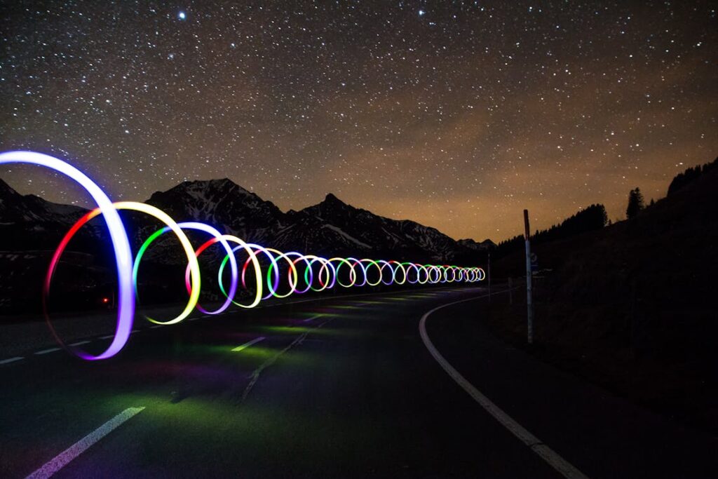 Free Enchanting long exposure of colorful light trails on a winding road beneath a star-filled sky. Stock Photo