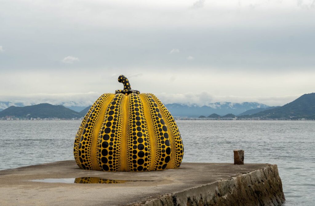 Free Yellow pumpkin sculpture by Yayoi Kusama on Naoshima Island pier, with sea and mountains in background. Stock Photo