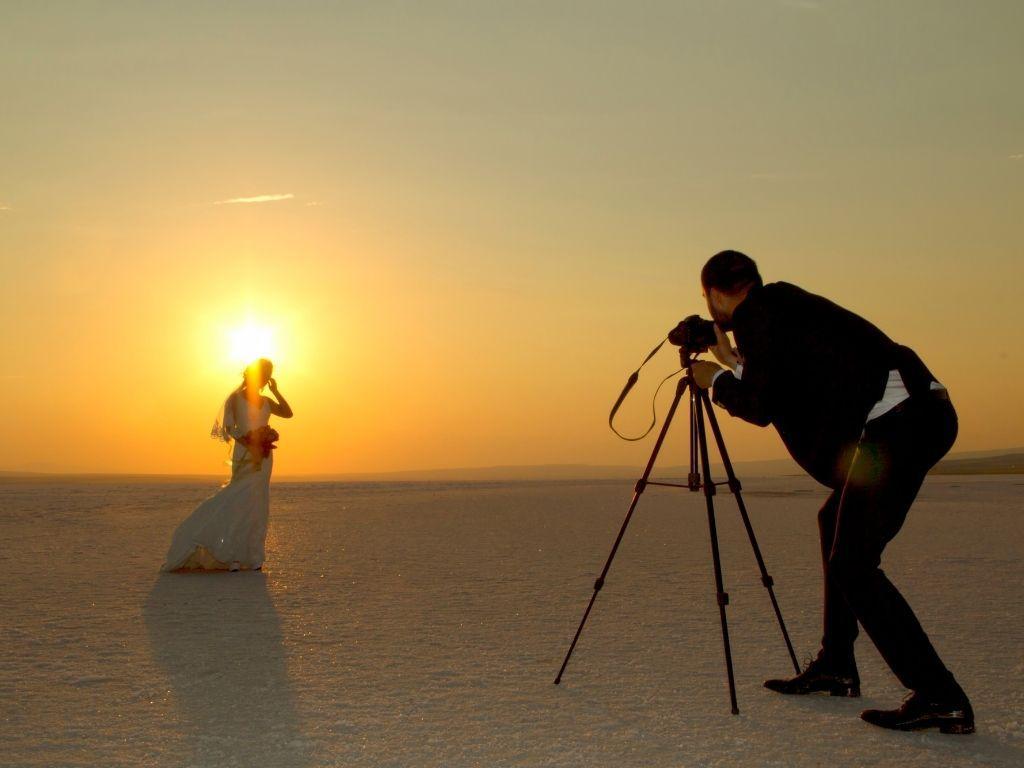 a man taking a photo of a bride holding a bouquet of red roses in a desert area while the sun is setting.